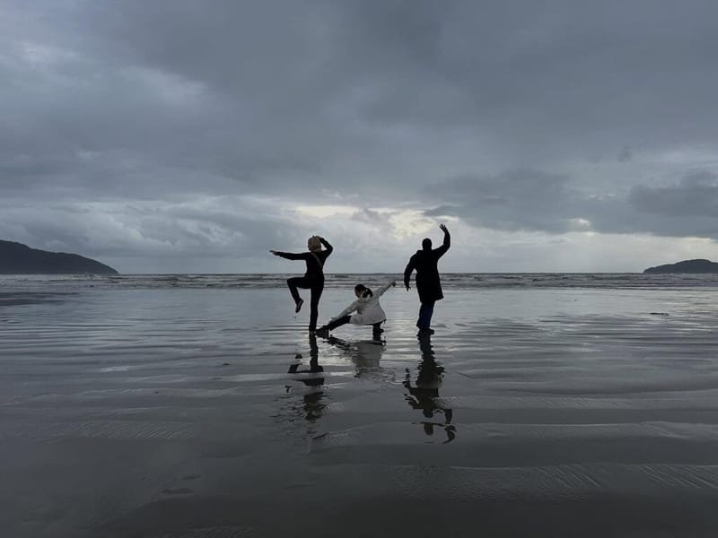 Three people striking playful poses on a wide wet beach under a dramatic overcast sky, small islands visible on the horizon