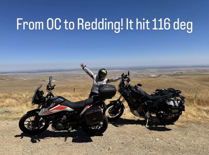 Motorcyclist standing between two adventure bikes on a hilltop overlook during a ride from Orange County to Redding, California, arms raised, dry golden hills stretching to the horizon