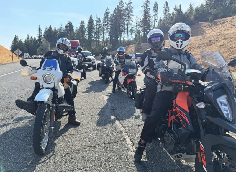 Group of motorcyclists in helmets and riding gear stopped on a highway pullout, pine-covered hills in the background