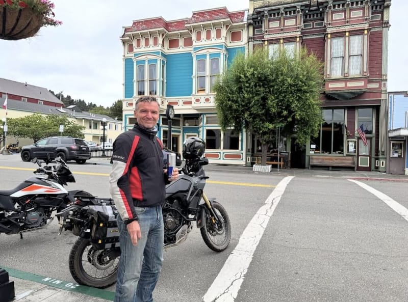 Man in motorcycle riding jacket standing in front of two adventure bikes and colorful Victorian-era storefronts on a small-town main street