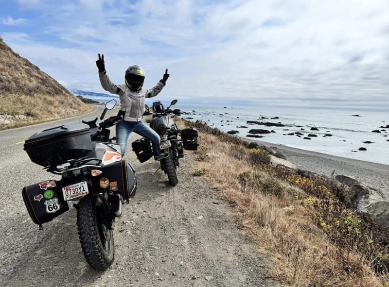 Rider in helmet standing on motorcycle footpegs with arms raised in a peace sign, two loaded adventure bikes parked on a coastal highway above a rocky shoreline