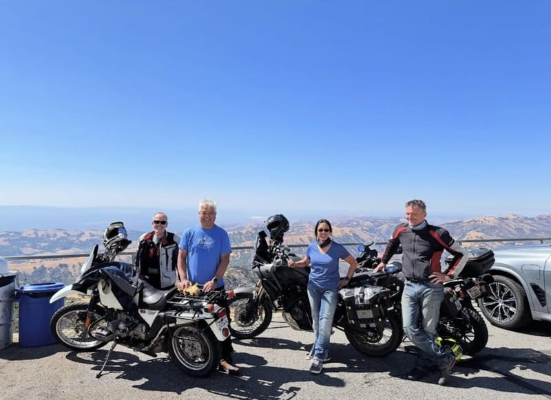 Four riders posing with adventure motorcycles at a hilltop overlook, rolling brown hills and blue sky stretching to the horizon
