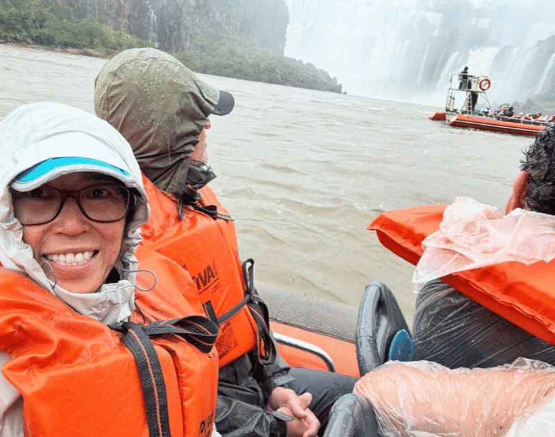 Andri Ayu smiling in an orange life jacket on a rubber raft at Iguazu Falls, with other passengers and mist-shrouded waterfalls in the background